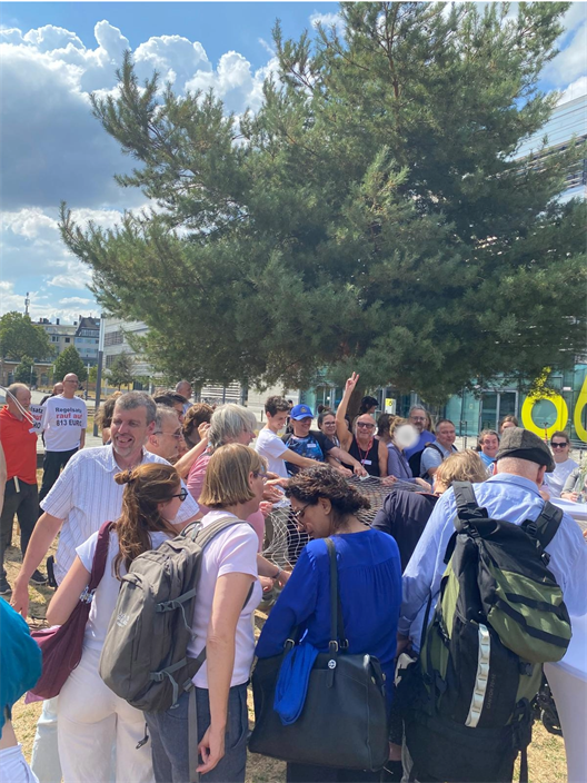 TMA 9 - Menschen stehen in einer großen Gruppe auf dem Campus der HSD und unterhalten sich. Im Hintergrund steht ein Baum. Die Menschen in der Mitte halten ein symbolisches Netz gemeinsam. Netz = Sicherung.